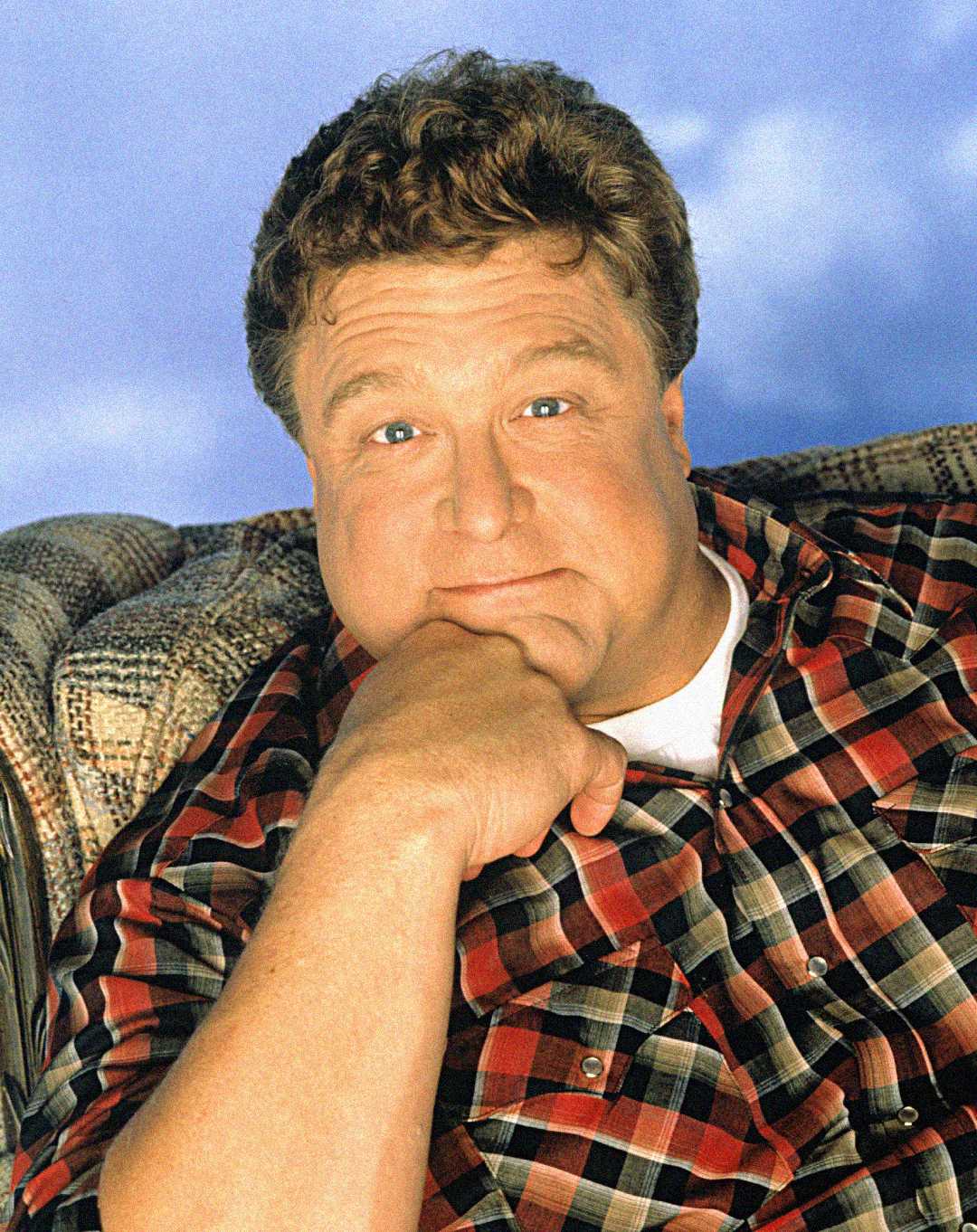 A man with short curly hair wearing a red and black plaid shirt sits on a patterned couch, resting his chin on his hand and looking at the camera with a gentle expression. The background is a soft blue.