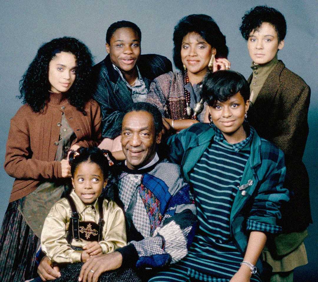 A group of seven people, including two men and five women, pose closely together and smile against a plain backdrop. The group appears to be a family, wearing colorful, casual clothing.