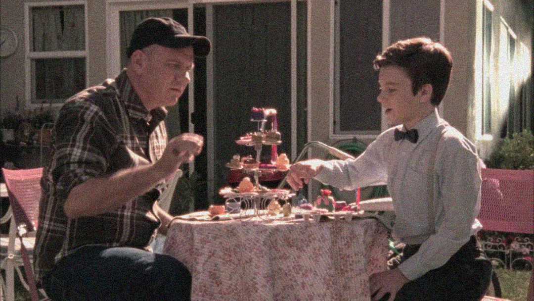 A man in a plaid shirt and cap sits at a small outdoor table with a boy in a dress shirt and bow tie, sharing snacks arranged on a tiered tray in a backyard setting.