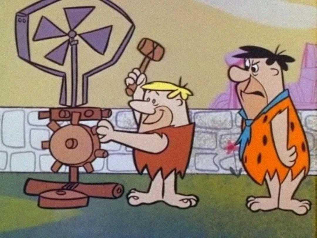 Two cartoon cavemen, one holding a hammer and working on a wooden contraption with gears and a windmill, while the other stands beside him with a surprised expression. Background shows a stone wall and pastel scenery.
