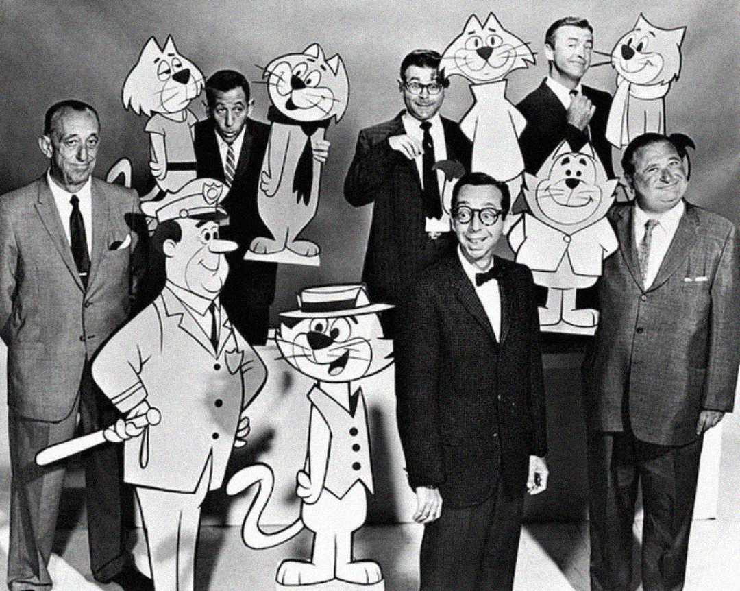 Five men in suits stand among large cutouts of animated "Top Cat" characters, including Top Cat himself, Officer Dibble, and others, against a plain backdrop. The mood is lively and celebratory.