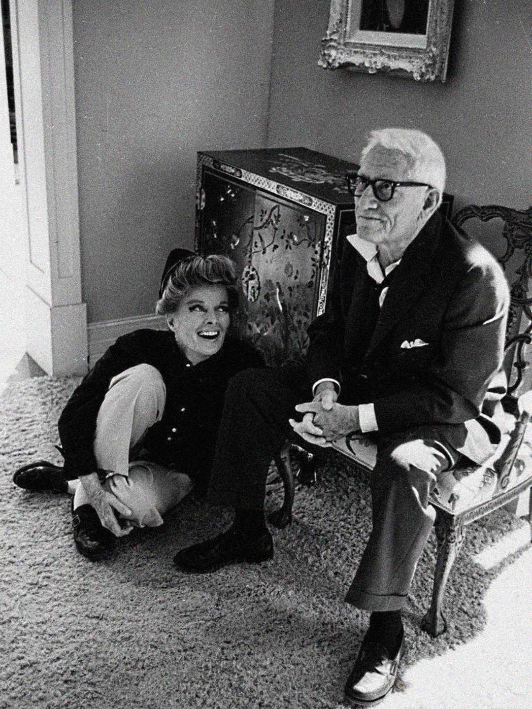 A woman sitting on the carpet, smiling up at an older man with glasses who is seated on a chair. They are indoors, near a decorative cabinet and a framed picture on the wall. The photo is in black and white.