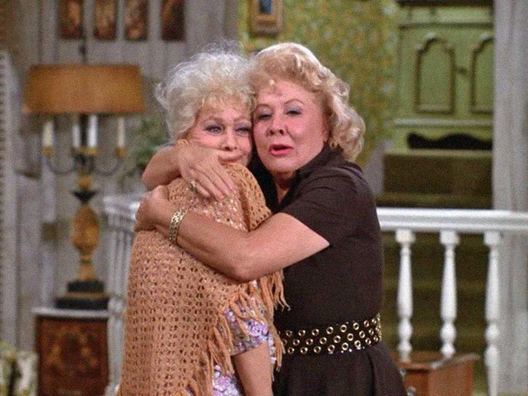 Two older women stand indoors, embracing warmly. One wears a light-colored crocheted shawl, while the other wears a dark dress with a studded belt. They appear emotional, standing in a cozy, vintage-style room.