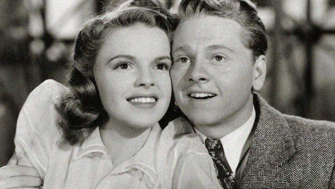A black-and-white photo of a smiling young woman and man, sitting close together. The woman has wavy hair and wears a light blouse; the man wears a patterned suit jacket and tie. Both look slightly upward, appearing happy.