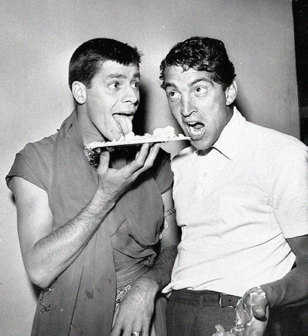 Two men in vintage clothing, one holding a pie and sticking out his tongue toward it, while the other stands beside him with his mouth open, as if about to eat. Both appear playful and amused in a black-and-white photo.