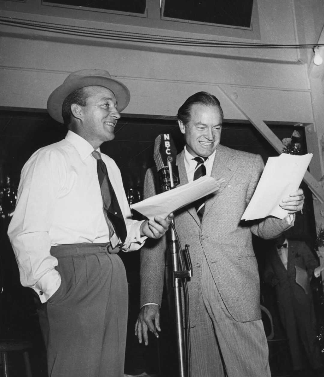 Two men in suits stand by an NBC microphone, smiling and holding papers, as if performing or rehearsing a radio show. One man wears a hat while the other is looking at the script.