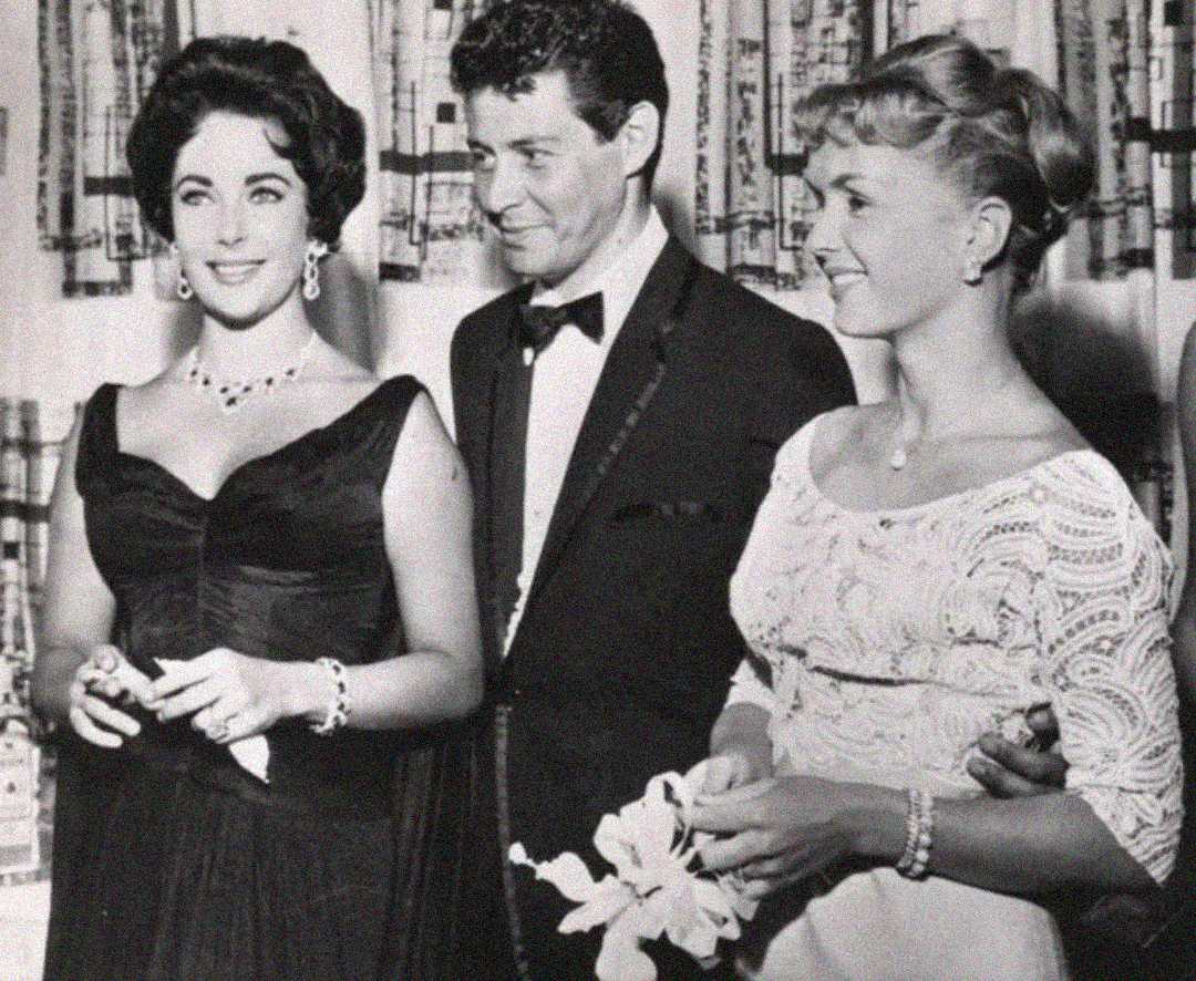 Three people in formal attire stand together, smiling for a photo. The woman on the left wears a dark gown and jewelry, the man in the center wears a tuxedo, and the woman on the right wears a light-colored dress and holds flowers.