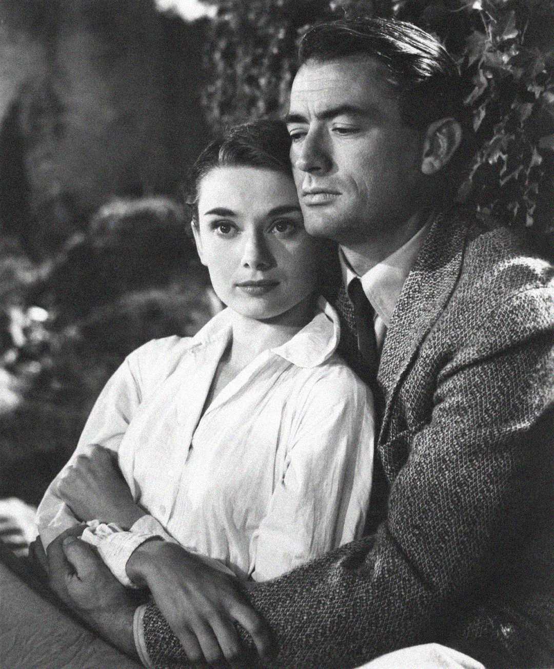 A black-and-white photo of a woman and a man sitting close together; the woman, wearing a white blouse, looks ahead while the man, in a suit jacket and tie, holds her and gazes thoughtfully into the distance.