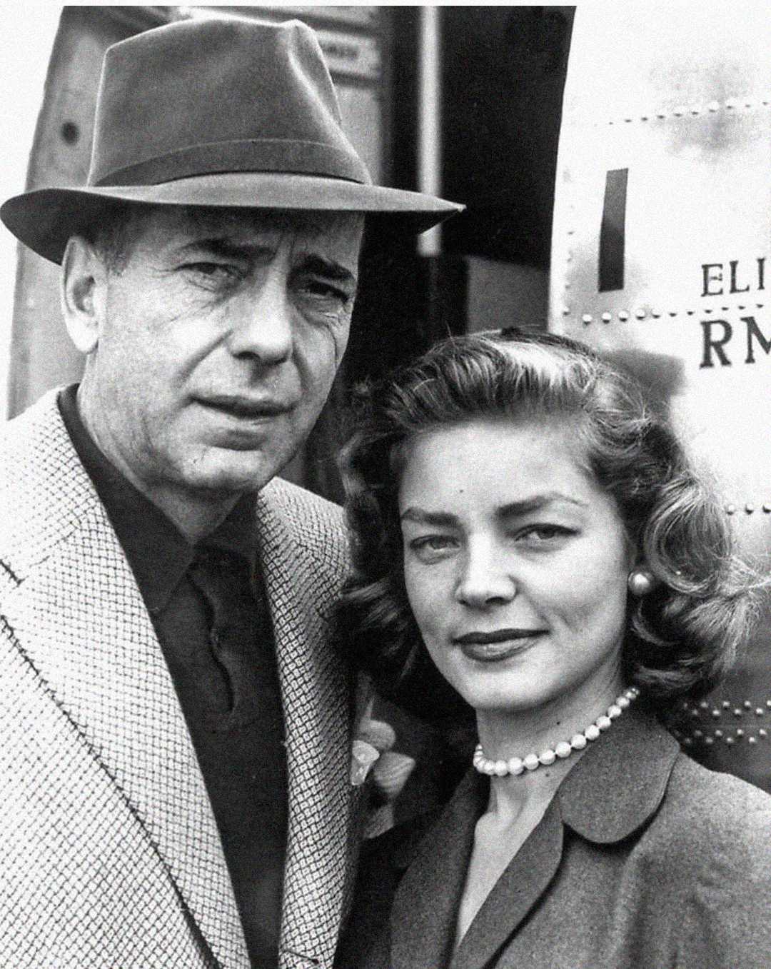 A man in a suit and fedora stands next to a woman wearing a pearl necklace and a blazer. Both are looking at the camera and smiling slightly. An airplane is visible in the background. The image is in black and white.