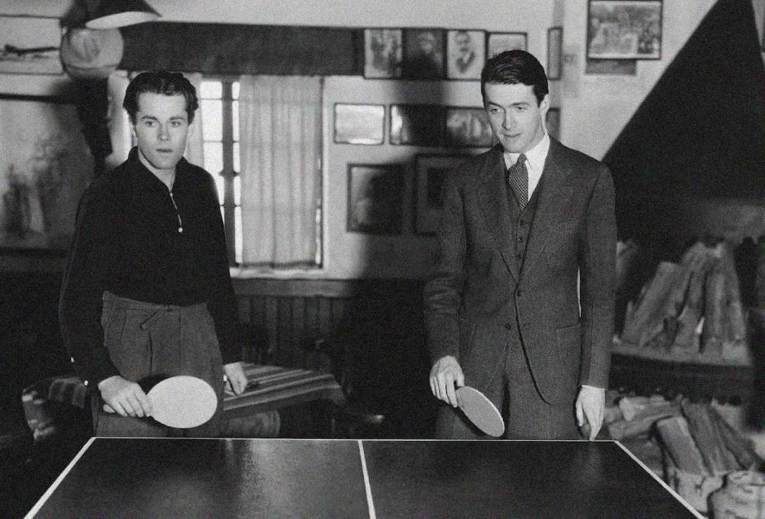 Two men, one in a sweater and the other in a suit, stand on either side of a ping pong table holding paddles. The room has framed pictures on the wall and a window with curtains in the background.