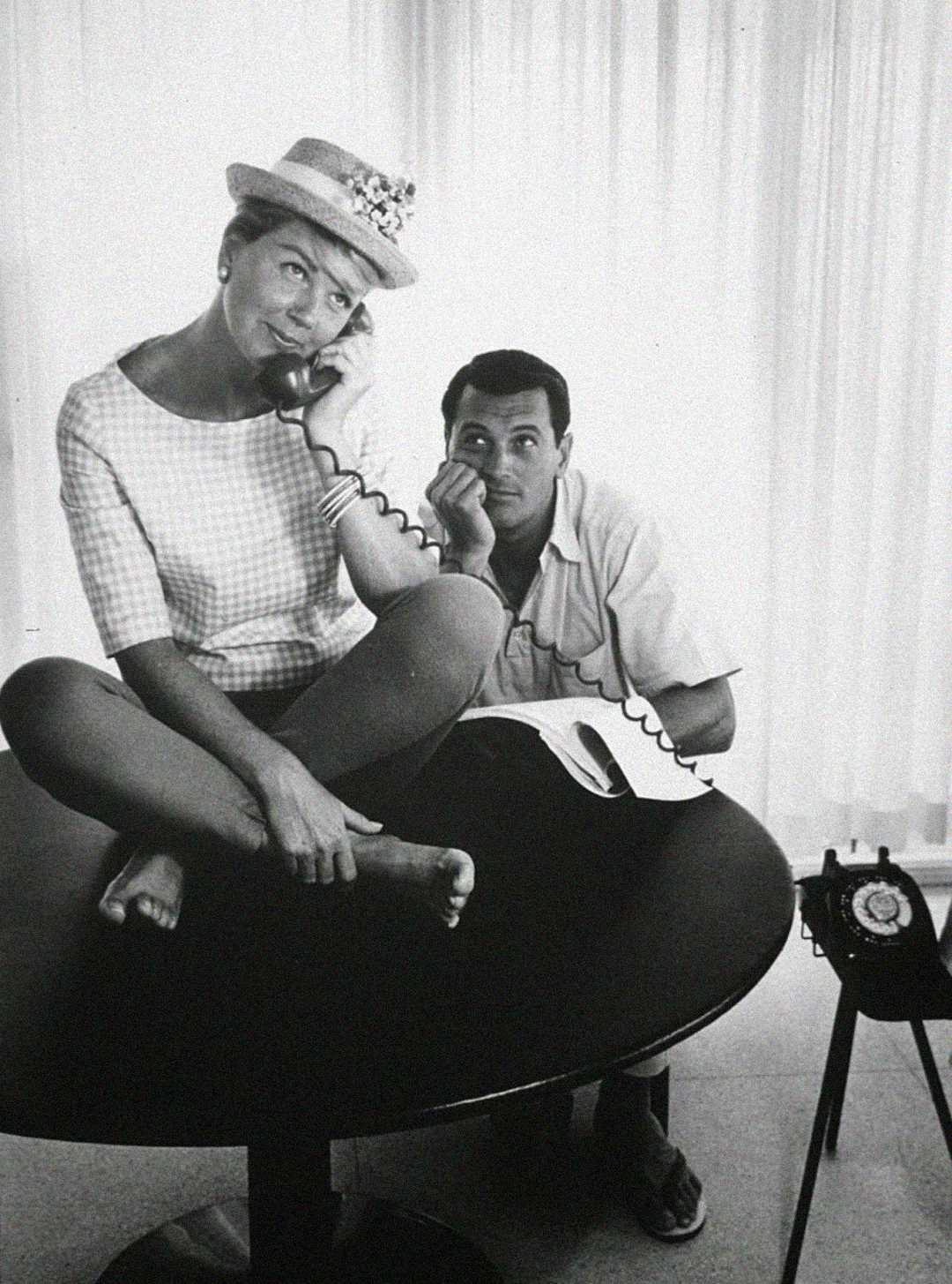 A woman sits cross-legged on a table, wearing a hat and holding a telephone receiver to her ear, while a man sits below her, looking up and listening. A rotary phone is on the table. Both appear thoughtful.