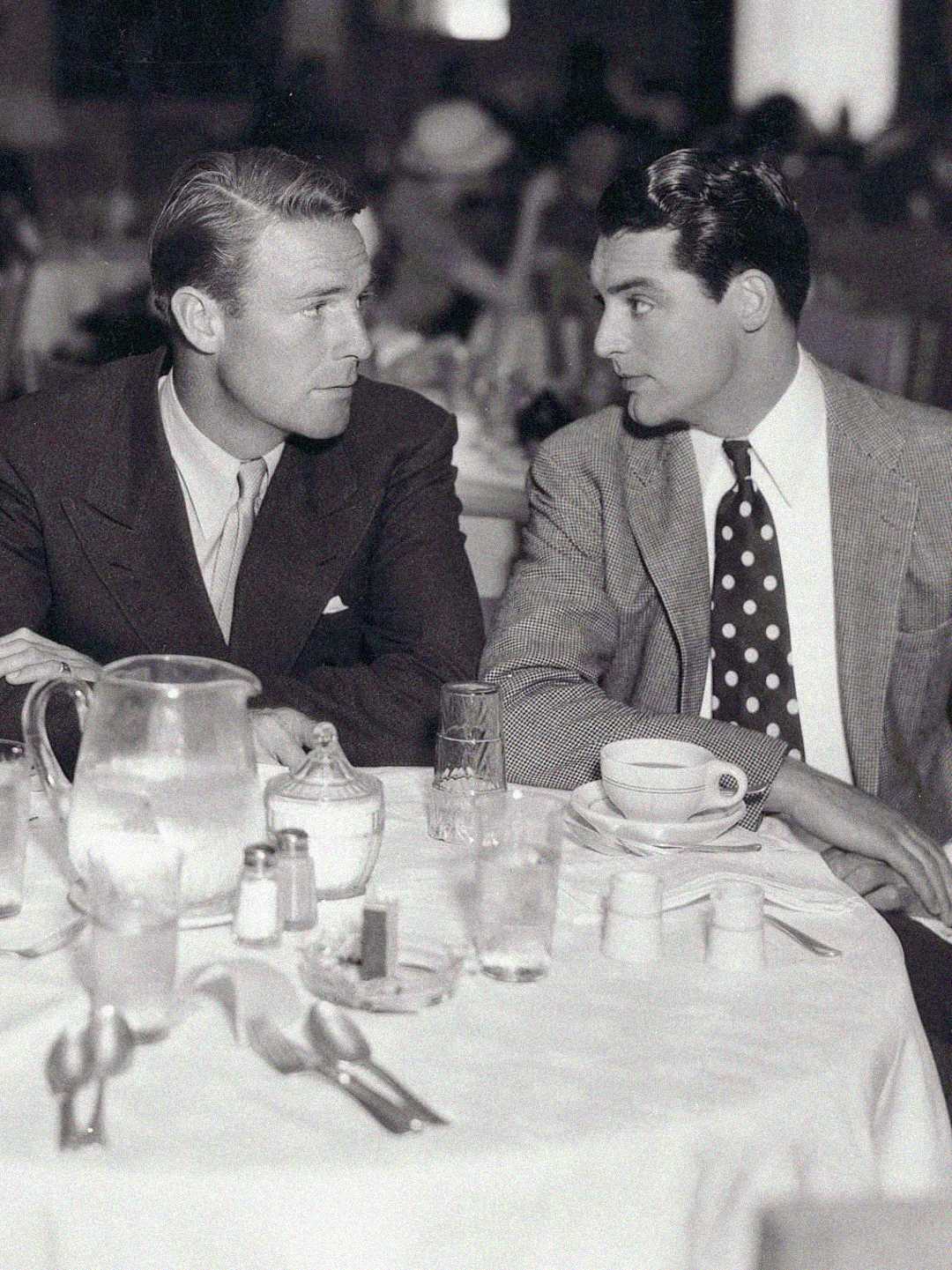 Two men in suits sit at a restaurant table set with a teapot, cups, a pitcher, and condiments, engaged in a serious conversation in a vintage black-and-white photo.