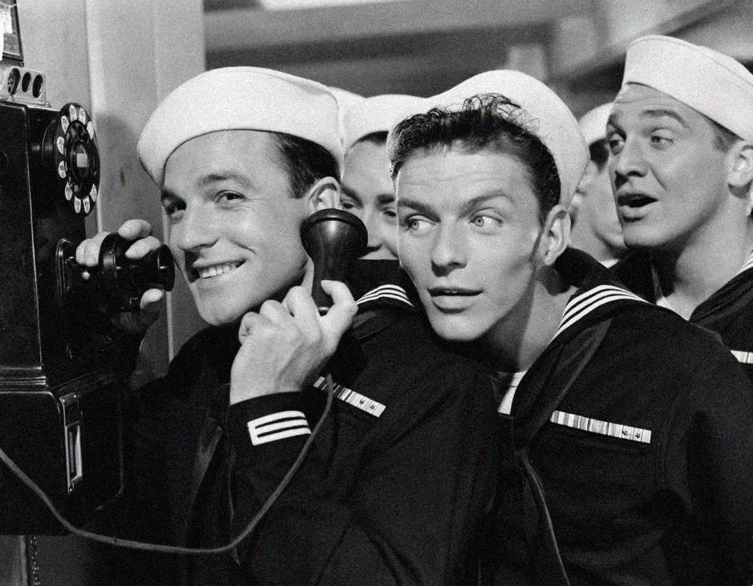 Three smiling sailors in uniform crowd around a rotary payphone, with one holding the receiver to his ear while the others eagerly listen in. The photo is black and white and has a cheerful, vintage feel.