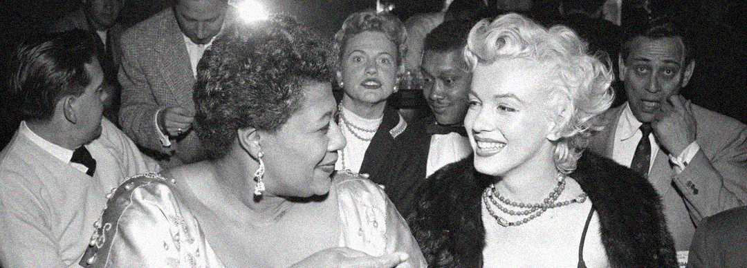 Two women in elegant attire smile and converse at a crowded event, surrounded by people; one has short curly hair and the other blonde waves, both wearing jewelry and glamorous outfits.