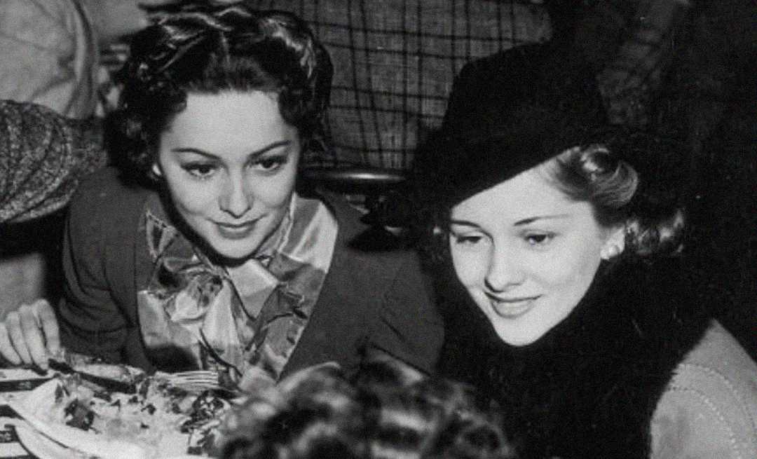 Two women in vintage clothing sit side by side at a table, smiling. One wears a hat and fur collar, the other has curled hair and a patterned scarf. The photo is black and white, suggesting it is from the early to mid-20th century.