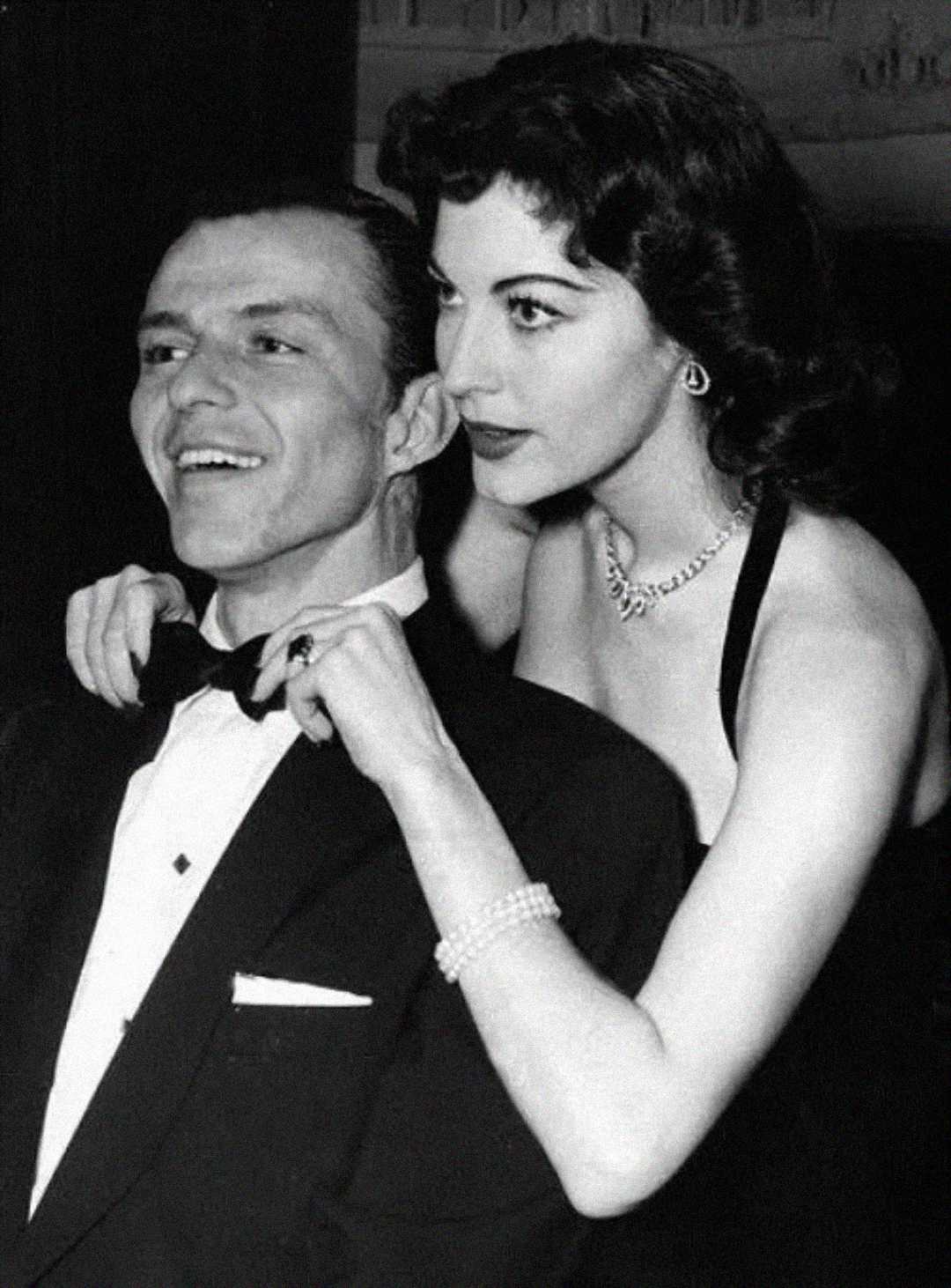 A smiling man in a tuxedo is having his bow tie adjusted by a woman in an elegant black dress and pearl jewelry. Both appear to be at a formal event or party.