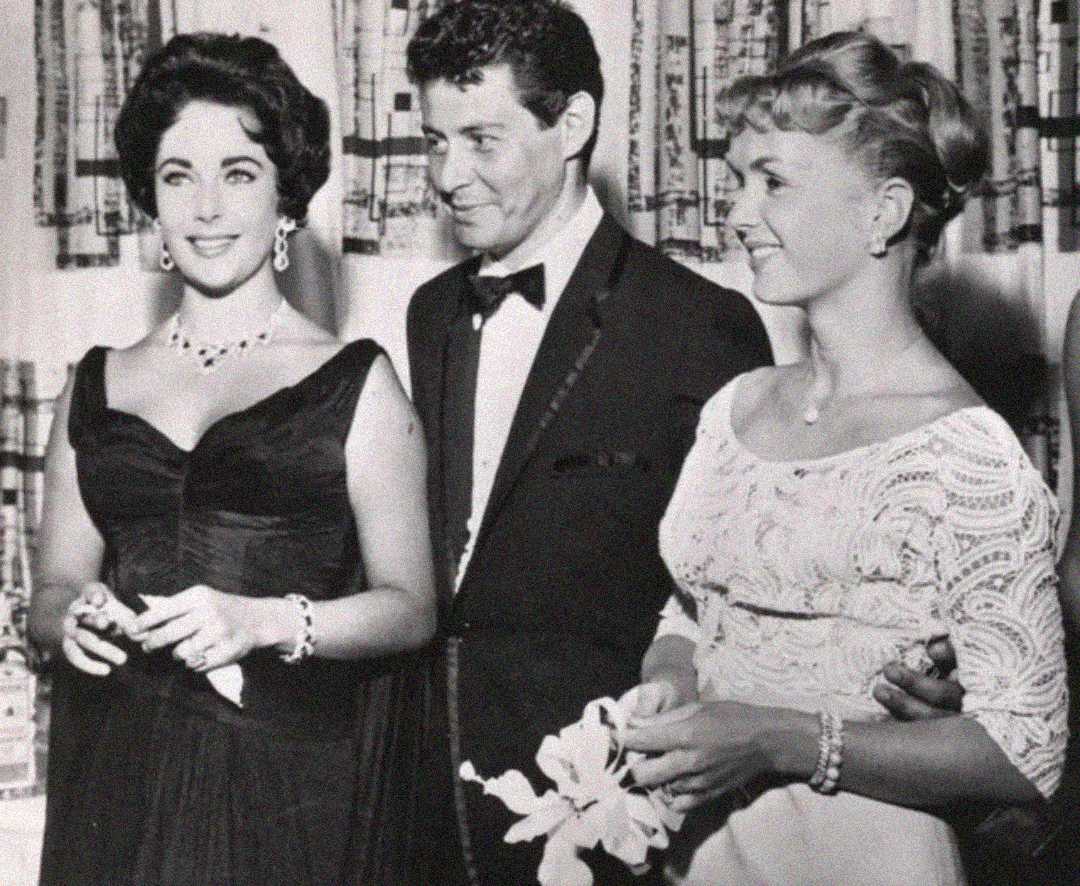 Three people dressed formally at an event; two women in elegant dresses stand on either side of a man in a tuxedo, all smiling. The setting appears to be indoors with patterned curtains in the background.