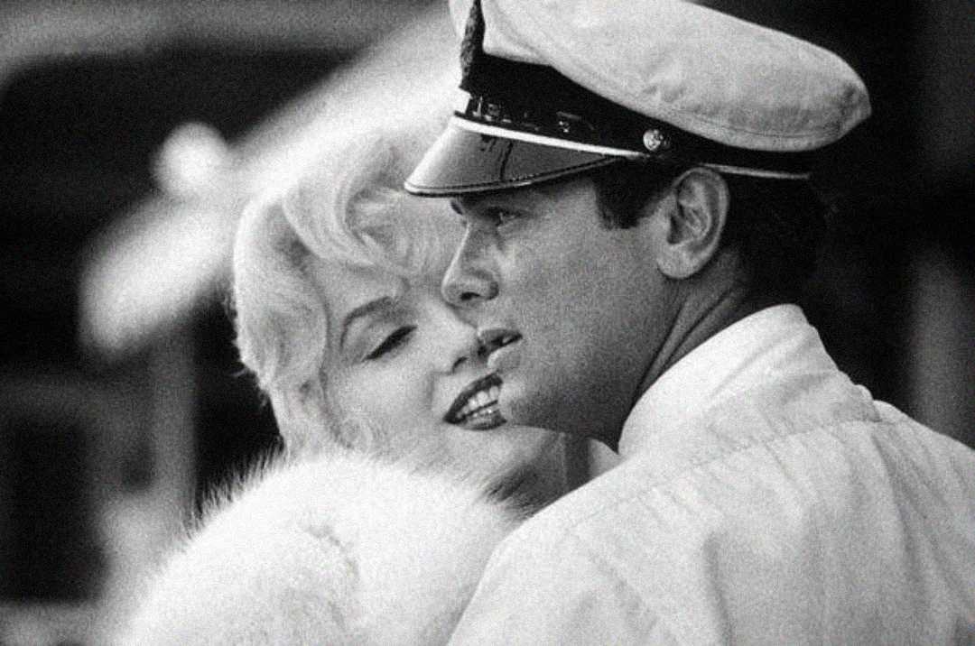A woman with blonde hair and a fur stole leans closely behind a man in a white naval officer's uniform and cap. The image is in black and white.