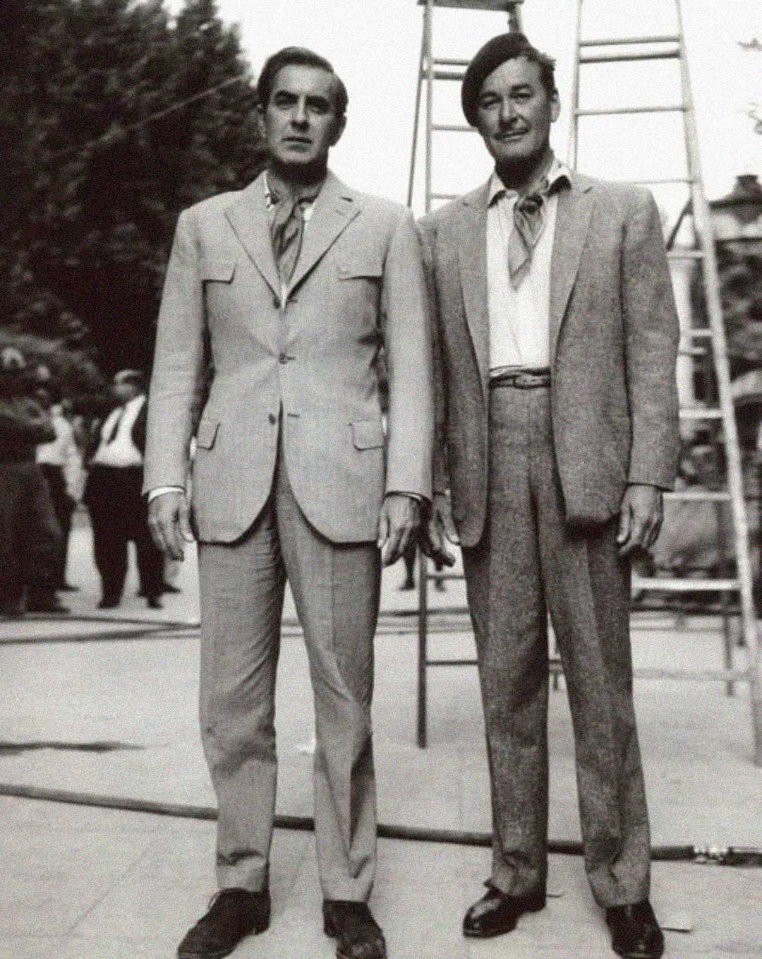 Two men in vintage suits stand side by side outdoors. One man wears a beret and a neck scarf. A ladder and several people are visible in the blurred background. The image appears to be black and white.