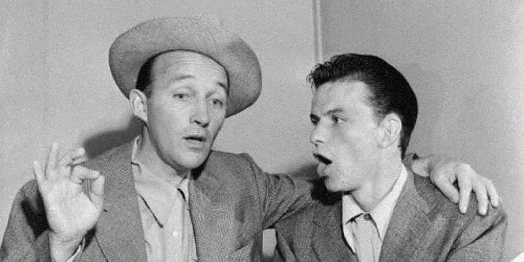 Two men in suits, one wearing a hat, pose together. The man on the left gestures an "OK" sign with his hand, while the man on the right looks at him with his mouth open as if singing or speaking.