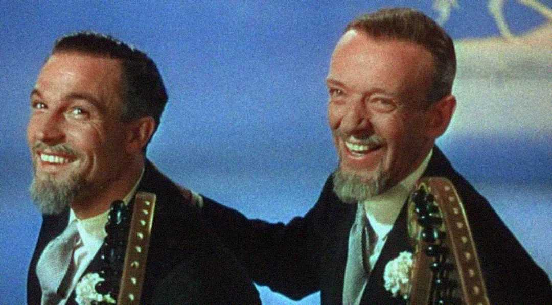 Two men in suits with gray goatees smile and laugh while holding guitars. They are dressed similarly, wearing white boutonnieres, and pose against a softly blurred blue background.