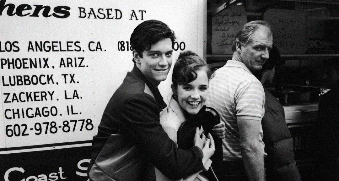 A young man hugs a smiling young woman in front of a food truck with city names and phone numbers listed; an older man stands nearby, looking over his shoulder. The photo is in black and white.