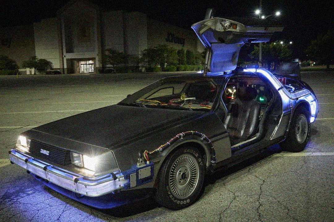 A silver DeLorean car with gull-wing doors open is parked in an empty lot at night. Blue accent lights illuminate the car, and wires are visible on its body, resembling the time machine from "Back to the Future.