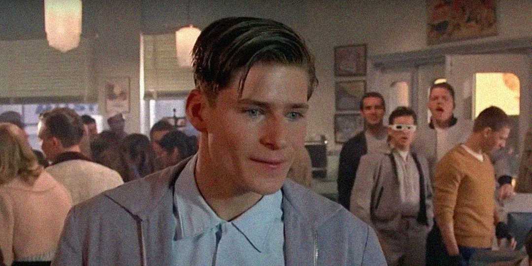 A young man with slicked-back hair and a light blue shirt stands in a crowded diner, looking slightly nervous. People in 1950s-style clothing are in the background, including one person wearing 3D glasses.