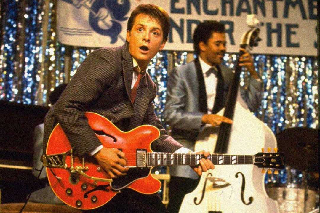 A young man in a suit plays a red electric guitar energetically on stage, with a surprised expression, while another musician plays an upright bass in the background. The stage is decorated with shiny silver streamers and a banner.