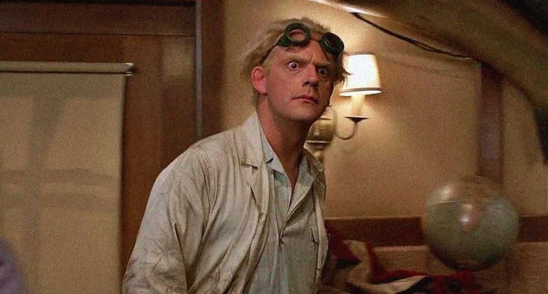 A man with wild white hair and goggles on his head wears a white lab coat and looks surprised. He stands indoors near a wall lamp and a globe. The setting appears to be a room with wooden paneling.