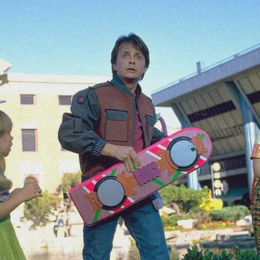 A young man in a futuristic vest holds a pink hoverboard, standing outside near a modern building, with a surprised expression on his face.
