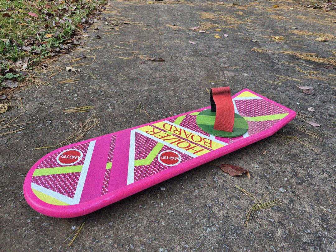 A pink hoverboard prop with yellow stripes, red foot strap, and "HOVER BOARD" text, lies on an outdoor path with grass, pine needles, and leaves around.