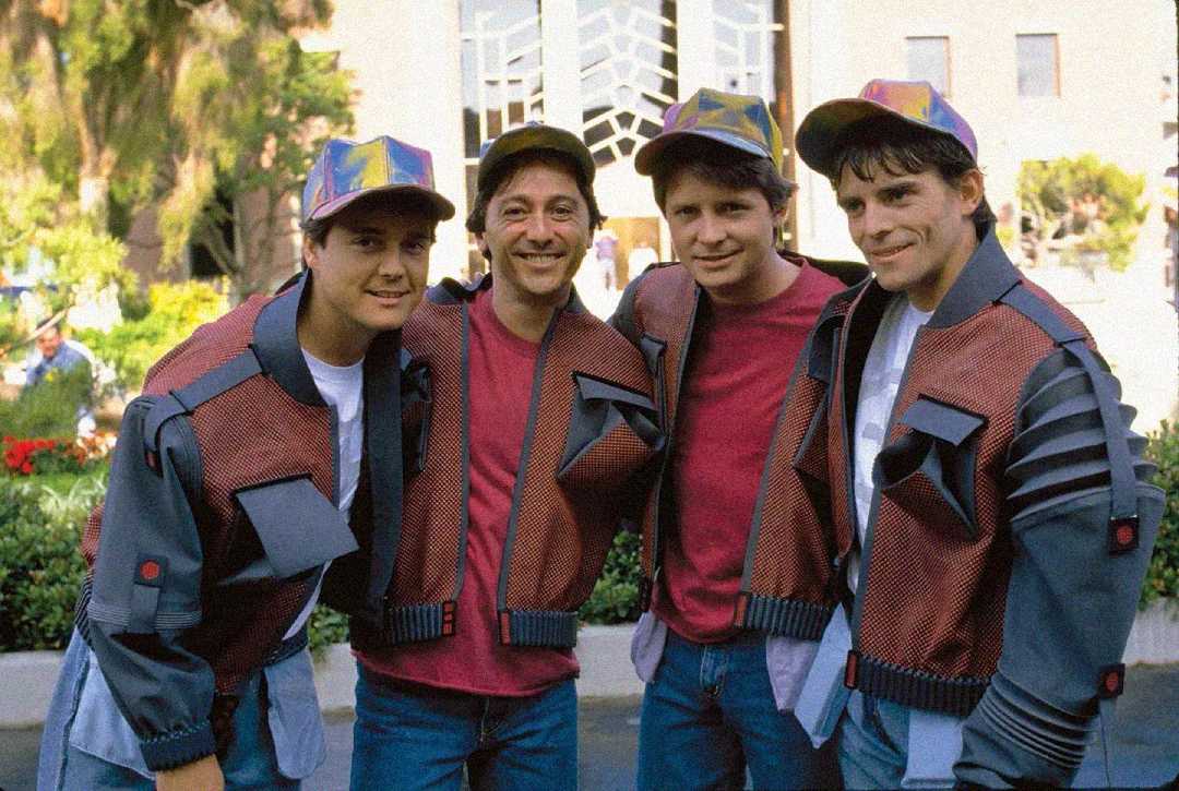 Four people stand outdoors, all wearing matching futuristic jackets and colorful, shiny caps. They are smiling, with greenery and a large building visible in the background. The mood is cheerful and casual.