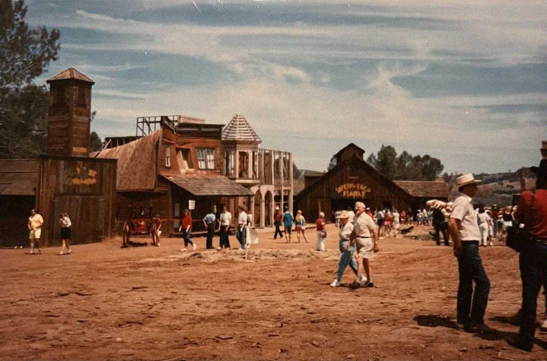 A group of people walk through a dusty, old western town with wooden buildings under a blue sky with wispy clouds; some people wear hats and casual clothes, evoking a theme park or movie set atmosphere.