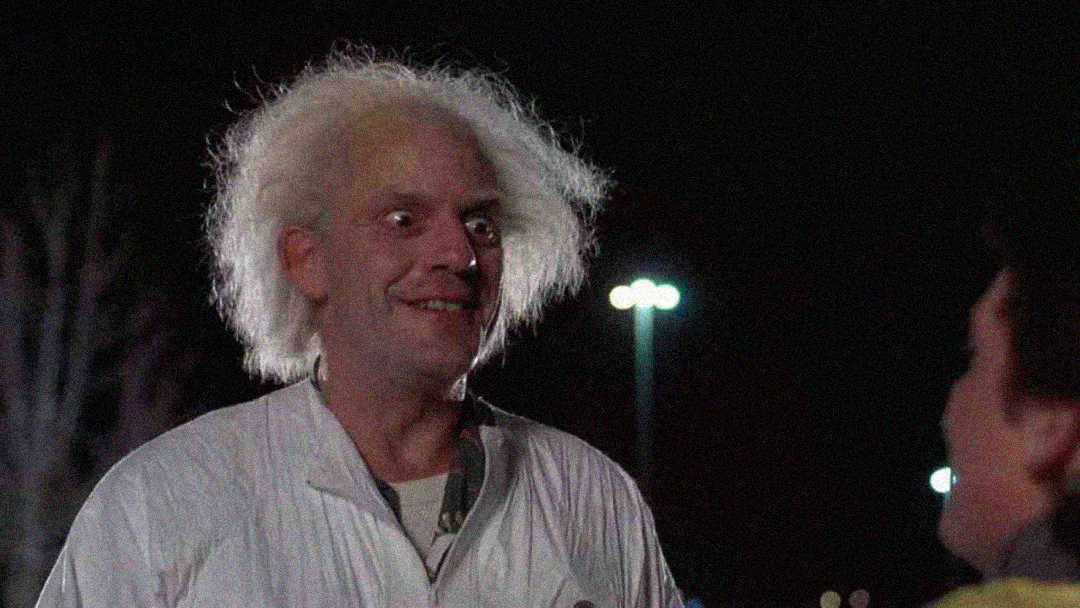 A man with wild, white hair and a surprised expression stands outdoors at night, illuminated by bright lights in the background. He is wearing a white coat and facing another person.