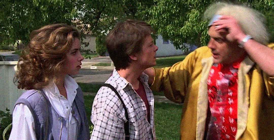 A young woman and a young man stand outdoors, looking at an older man with messy white hair and a yellow coat, who appears to be speaking urgently to them. Trees and houses are visible in the background.
