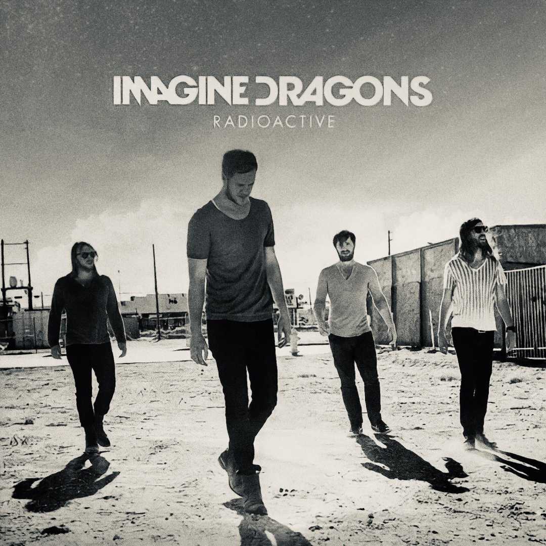 Four men walk on a sunlit, sandy ground, casting long shadows. The words "IMAGINE DRAGONS RADIOACTIVE" appear at the top of the black-and-white image. Industrial structures are visible in the background.