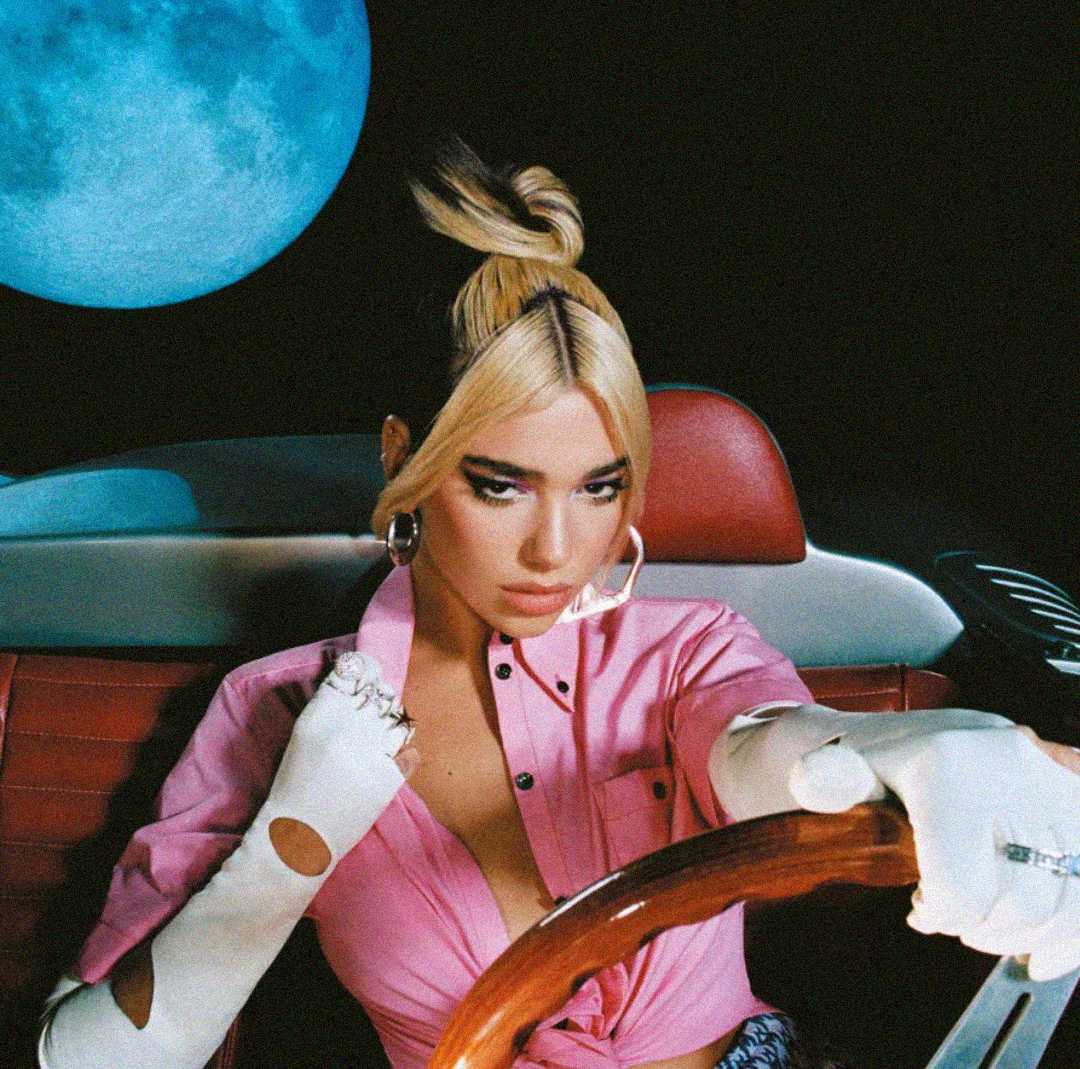 A woman with blonde hair in a high bun sits in a red car seat, wearing a pink top and white gloves. She holds a steering wheel and looks forward, with a large blue moon in the dark sky behind her.