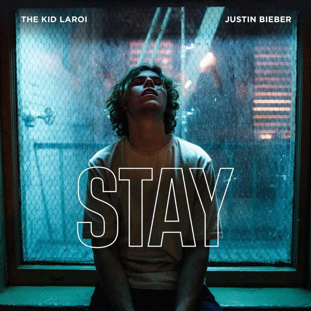 A young man sits in front of a large window with blue lighting, looking up. The words "THE KID LAROI," "JUSTIN BIEBER," and "STAY" are overlaid on the image in white text.