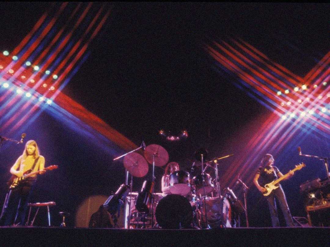 A rock band performs on a dark stage with colorful, crisscrossing lights overhead. Three musicians play guitar, drums, and bass, surrounded by musical equipment.