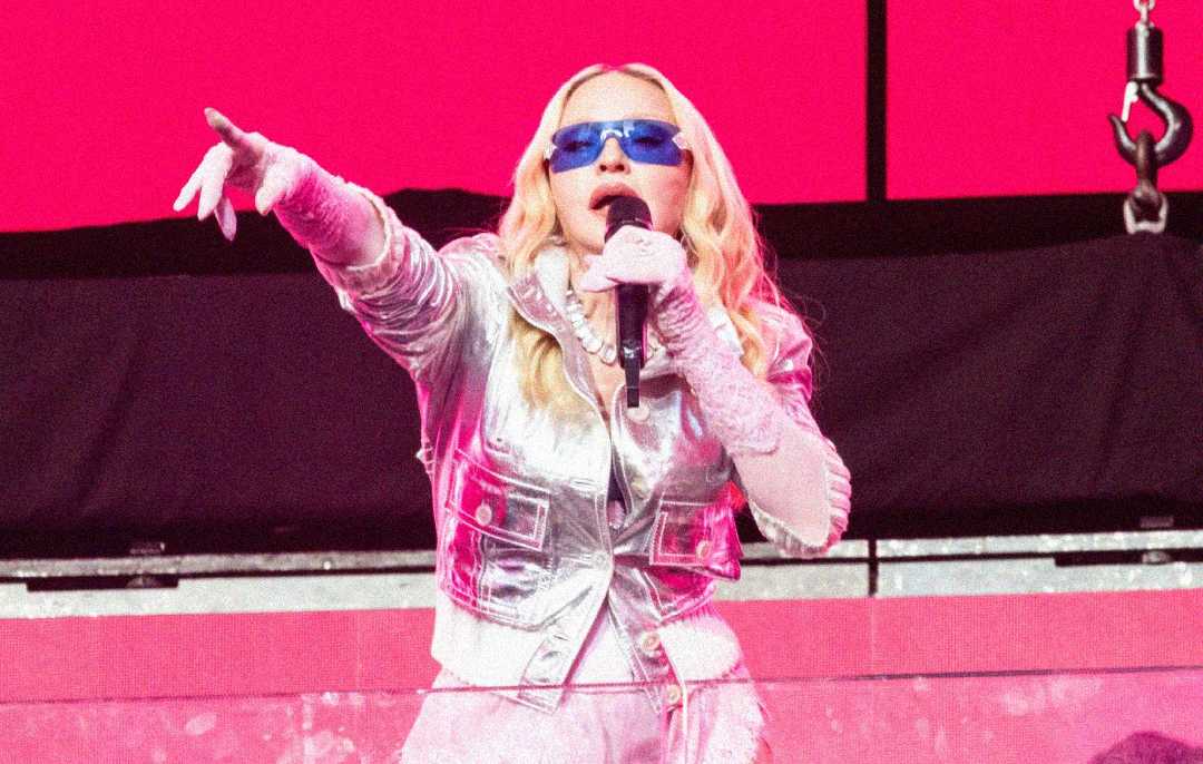 A performer in a shiny silver outfit and blue sunglasses sings into a microphone on stage, with a bright pink background behind them.