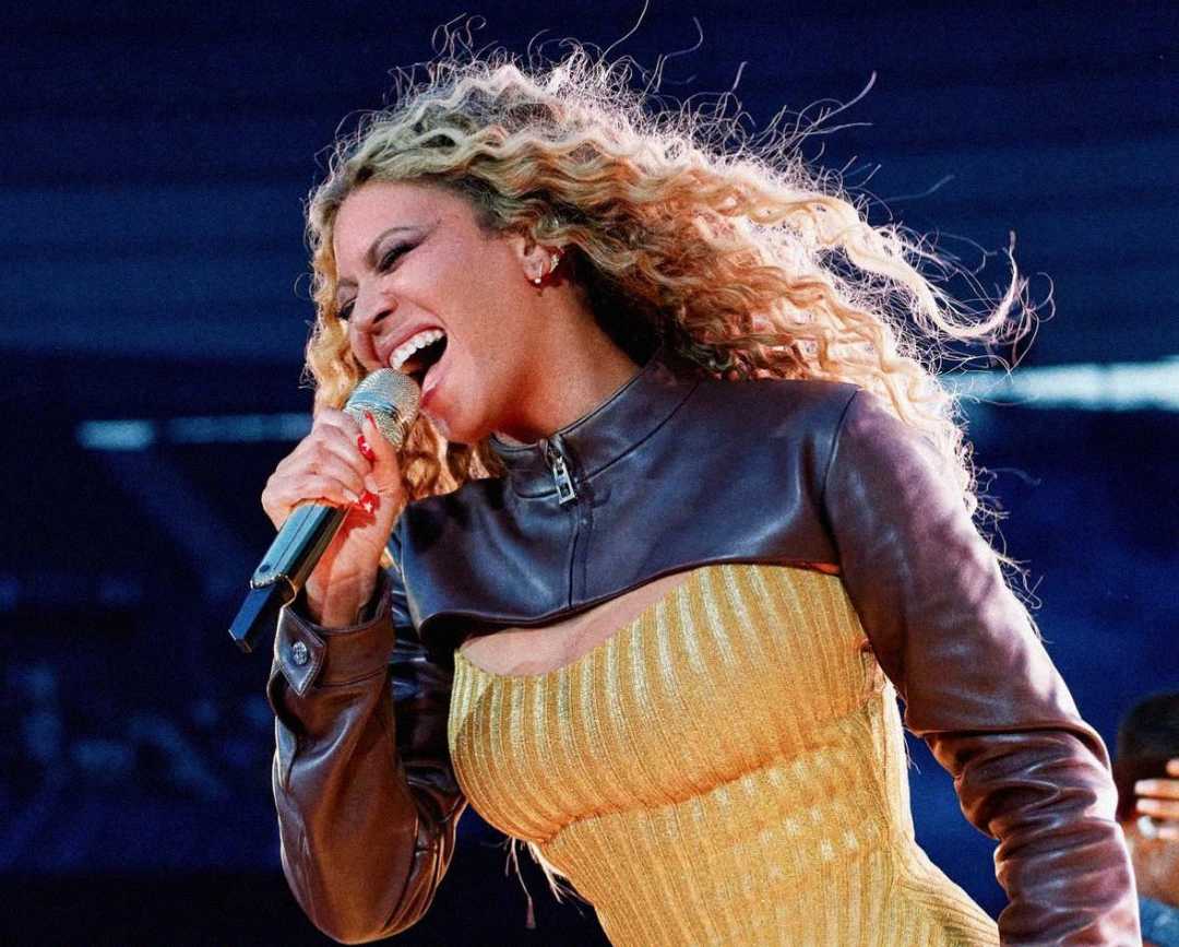 A woman with long curly hair sings passionately into a microphone. She wears a yellow dress with a brown leather top and performs on stage with a blurred background.