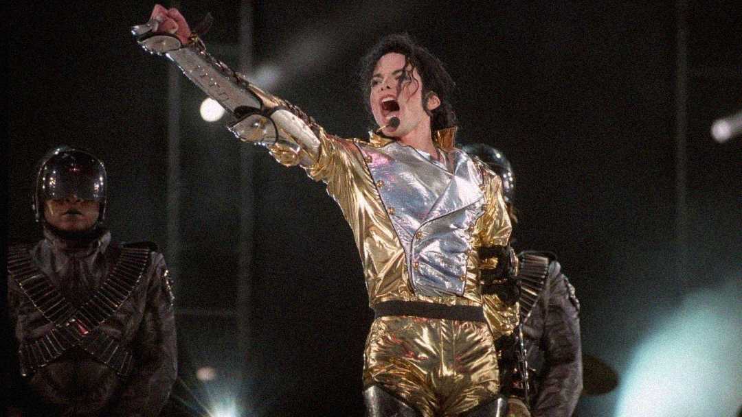 A performer in a shiny gold and silver futuristic outfit strikes a powerful pose on stage, with dancers in metallic costumes and helmets in the background, under dramatic lighting.