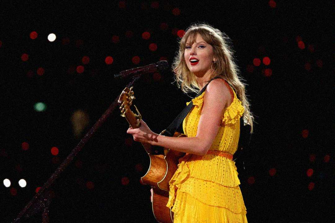 A woman in a bright yellow dress plays an acoustic guitar and sings into a microphone on stage, with soft red and white lights glowing in the dark background.
