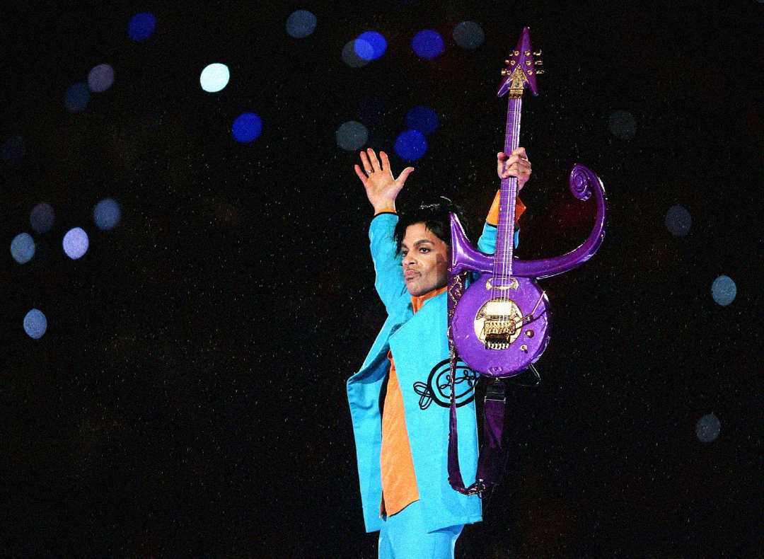 A performer wearing a blue suit with an orange shirt holds a purple guitar and raises one arm, standing on stage against a background of colorful, blurred lights.
