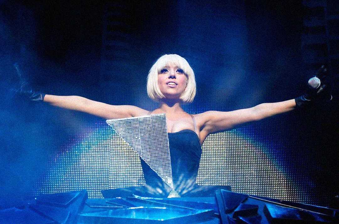 A performer with a platinum blonde bob and black gloves stands on stage, arms outstretched, wearing a dramatic geometric costume with a sparkling triangular front piece against a blue-lit backdrop.