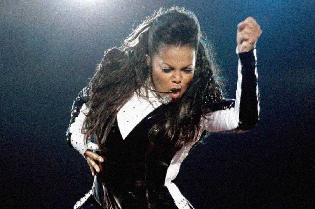 A woman with long hair performs energetically on stage, wearing a black and white outfit. She raises her right arm with a clenched fist and has an intense, focused expression on her face.
