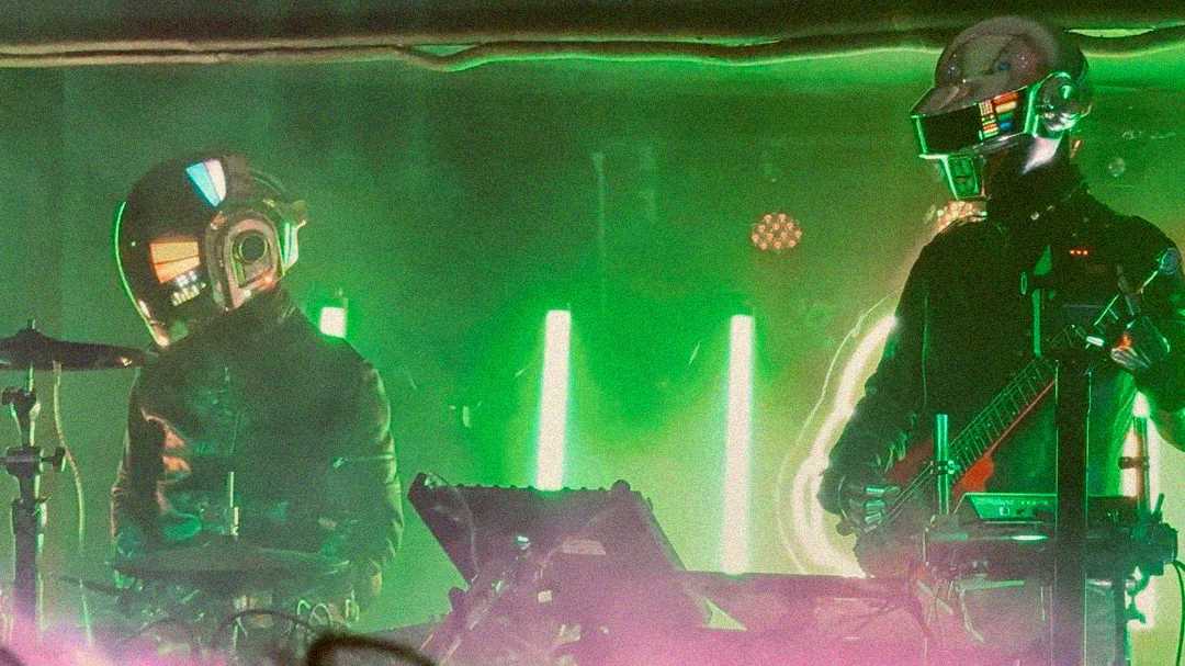 Two performers wearing futuristic, illuminated helmets and dark outfits play electronic instruments on a neon-lit stage with green and pink lighting effects.