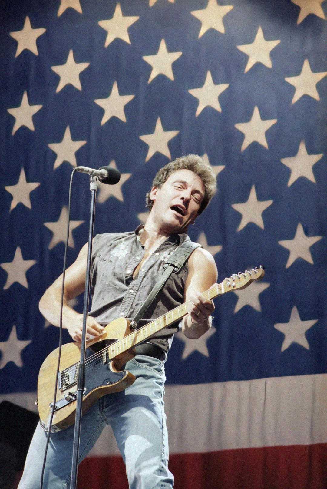 A man passionately plays an electric guitar and sings into a microphone on stage, with a large American flag featuring white stars and stripes as the backdrop.