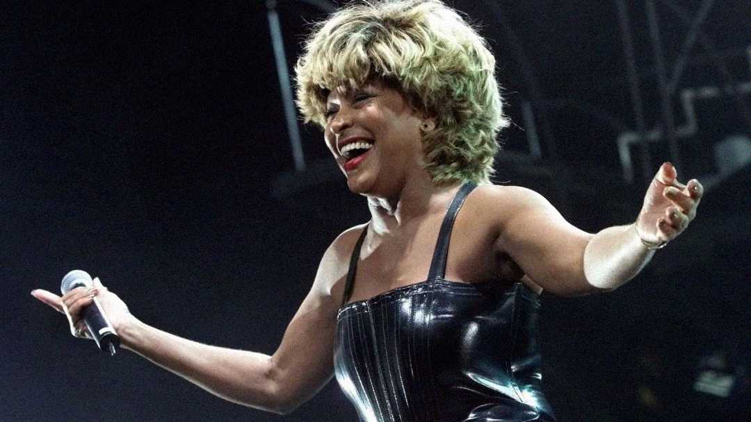 A woman with short, blonde hair wearing a shiny black dress smiles and holds a microphone on stage with her arms open wide, under bright stage lights.